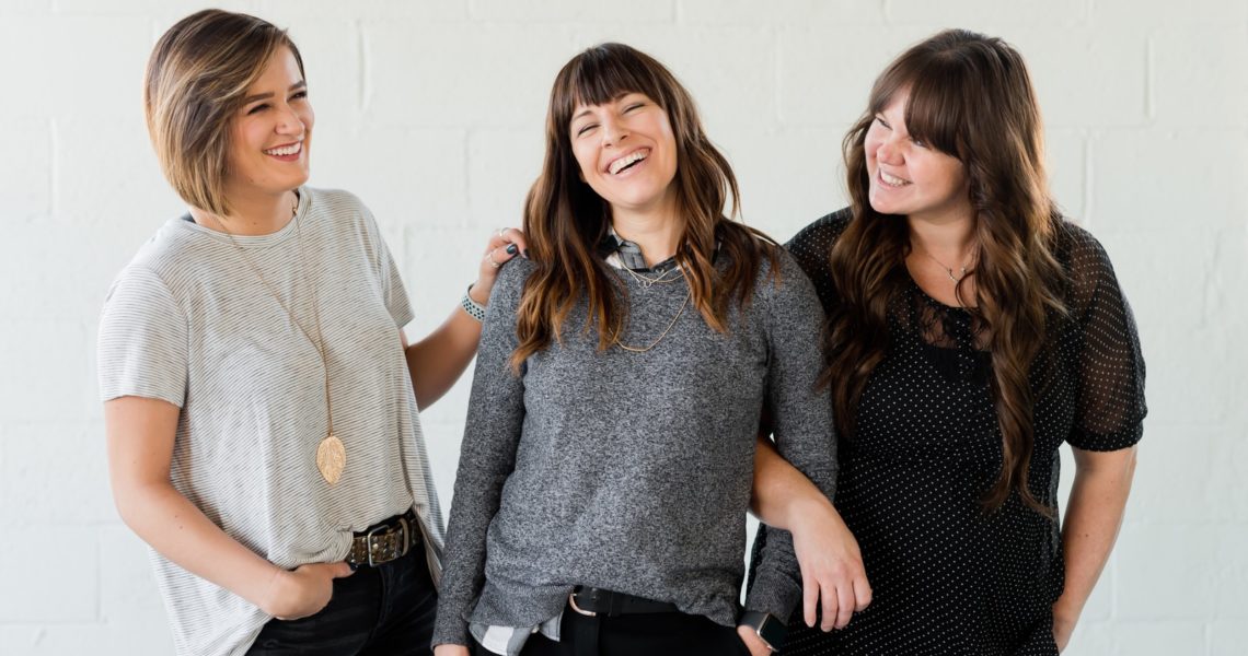 3 women smiling and standing beside white wall